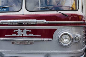 A vintage bus with a distinctive front design, featuring an emblem and retro headlights. The bus has a two-toned color scheme with maroon and silver hues and chrome accents. Visible portions of the interior show the steering wheel and a partial view of a person inside.