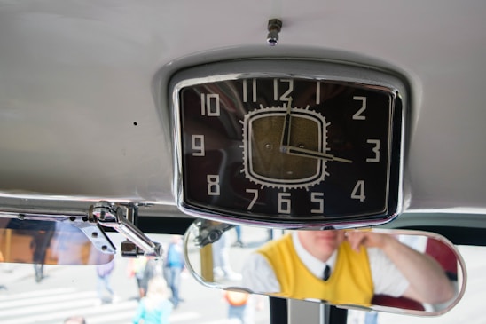 A retro-style clock with an analog face is mounted on the ceiling. The time is approximately 9:15. Below the clock, a vehicle's rear-view mirror reflects a person wearing a yellow vest and a white shirt with a black tie. The scene appears to be inside a vehicle with people visible outside through the windows.