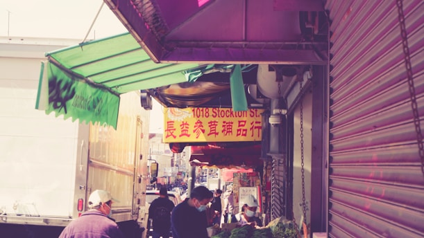 A bustling street market with people shopping and browsing stalls. Bright signage with Chinese characters is visible, and there are various awnings and a rolled-up shutter on the right. The area appears lively and busy.