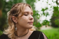 A person with closed eyes and glittering star shapes on their cheek stands outdoors. They have a peaceful expression, with light makeup and jewelry, including a nose ring and a mushroom-shaped earring. The background is lush and green, suggesting a natural setting.