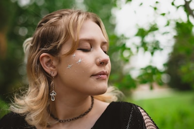 A person with closed eyes and glittering star shapes on their cheek stands outdoors. They have a peaceful expression, with light makeup and jewelry, including a nose ring and a mushroom-shaped earring. The background is lush and green, suggesting a natural setting.