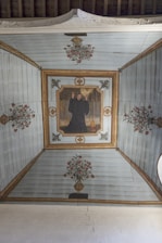 A painted ceiling depicting a person in a dark robe raising hands, surrounded by floral motifs and decorative patterns on a wooden surface. The central figure is framed by an ornate border, and there are symmetrical flower vases at each end, creating a harmonious and balanced composition.