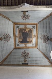 A painted ceiling depicting a person in a dark robe raising hands, surrounded by floral motifs and decorative patterns on a wooden surface. The central figure is framed by an ornate border, and there are symmetrical flower vases at each end, creating a harmonious and balanced composition.