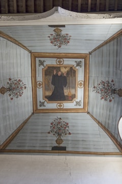 A painted ceiling depicting a person in a dark robe raising hands, surrounded by floral motifs and decorative patterns on a wooden surface. The central figure is framed by an ornate border, and there are symmetrical flower vases at each end, creating a harmonious and balanced composition.