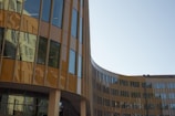 A modern, curved office building with large reflective glass windows and brown paneling, reflecting the surrounding environment. The structure features a sleek, contemporary design with clean lines and an open, airy façade.