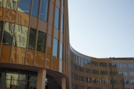 A modern, curved office building with large reflective glass windows and brown paneling, reflecting the surrounding environment. The structure features a sleek, contemporary design with clean lines and an open, airy façade.