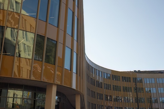 A modern, curved office building with large reflective glass windows and brown paneling, reflecting the surrounding environment. The structure features a sleek, contemporary design with clean lines and an open, airy façade.