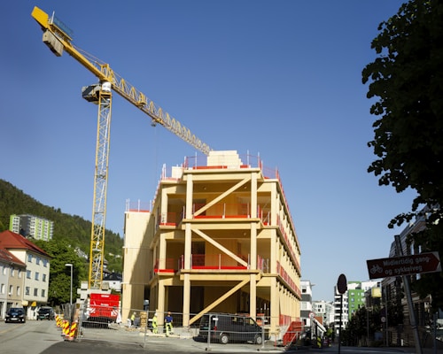 A construction site featuring a partially built multi-story wooden building with red accents is visible. A large yellow tower crane stands adjacent to the building, reaching into a clear blue sky. Various construction workers are seen working around the site, which is enclosed with fencing. Trees and other urban buildings surround the area, creating a blend of nature and urban development.