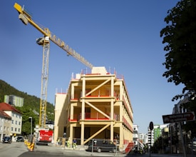 A construction site featuring a partially built multi-story wooden building with red accents is visible. A large yellow tower crane stands adjacent to the building, reaching into a clear blue sky. Various construction workers are seen working around the site, which is enclosed with fencing. Trees and other urban buildings surround the area, creating a blend of nature and urban development.