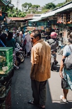 A busy outdoor market scene with people walking through a narrow pathway. Stalls line the street, which appear to be selling various items. A man in a patterned orange shirt is prominently in the foreground, while other people are going about their activities. Motorcycles are parked along the sides, and there is a feeling of crowdedness.