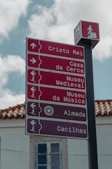 A tall signpost with several red directional signs pointing to various locations, including Cristo Rei and Casa da Cerca. Each sign has icons indicating walking directions. A smaller overhead sign shows a graphic of a person walking.