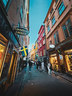 A narrow, vibrant street lined with colorful buildings in shades of red, orange, and yellow. Pedestrians, including families and individuals, walk along the cobblestone path. Shops with wooden facades display signs, some featuring Swedish flags, suggesting a European setting. The sky is clear and blue, enhancing the lively and bustling atmosphere.