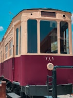 A vintage, maroon and beige tram is set against a clear blue sky. The tram has a rounded front with large windows, and the word 'Heliopolis' is visible on a sign above the windows. The tram has additional details like a circular ventilation cover and metal railings around the base.