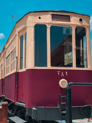 A vintage, maroon and beige tram is set against a clear blue sky. The tram has a rounded front with large windows, and the word 'Heliopolis' is visible on a sign above the windows. The tram has additional details like a circular ventilation cover and metal railings around the base.