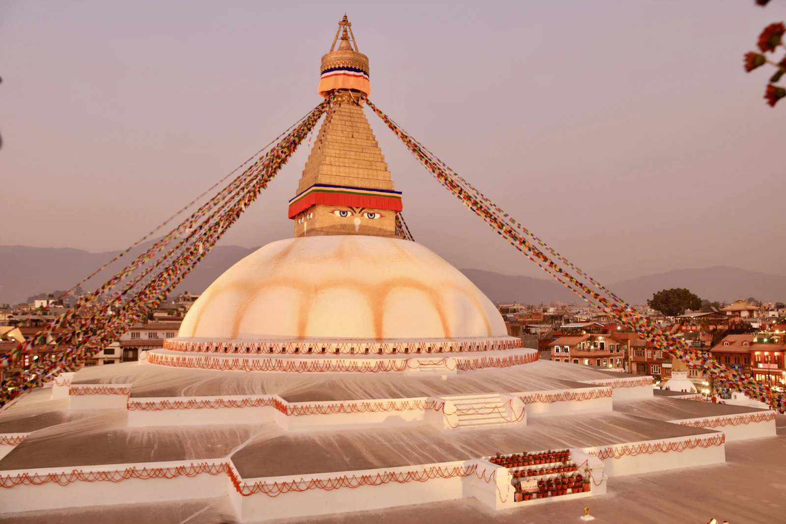 Boudhanath stupa at dusk in Kathmandu, its white dome, gilded tower and prayer flags seen from a rooftop under a pink sky