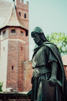 A bronze statue of a knight in detailed medieval armor stands prominently in front of a historic brick tower. The knight is wearing a helmet and a long robe, displaying a stoic posture. The background features a blurred view of trees and a clear sky, adding contrast to the dark tones of the statue.