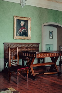 A room with classic wooden furniture, including an ornate chair and table. The green walls are adorned with a large, framed portrait of a historical figure wearing a luxurious garment. The room features intricate architectural details and a smaller artwork in the background.