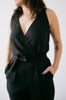 A person is wearing a sleeveless black jumpsuit with a V-neckline and a waist tie. The individual has tattoos on both arms and blue nail polish is visible on the right hand, which is placed in the pocket of the jumpsuit. The person is looking to the side against a plain white background.