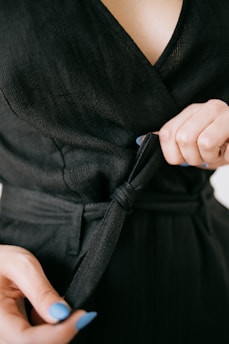 A person is adjusting a black fabric belt on clothing. The close-up view emphasizes the texture of the fabric and the details of the hands with blue painted nails.