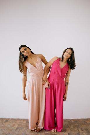 Two people are standing close together, each wearing a sleeveless jumpsuit. One person is in a light pink outfit and the other in a bright pink one. They are leaning slightly towards each other with relaxed postures against a plain white background on a wooden floor.