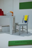 The image features a minimalist installation with a light gray chair draped with a yellow and blue cloth. A white, rectangular table is present, with geometric shapes including a red object, an orange cube, and a white polygon. A green and white two-toned wall decoration is visible, along with a green plank on the floor with black text 'CHRONONHOTONTHO 1734'. The setting appears to be an art or design exhibition.