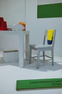 The image features a minimalist installation with a light gray chair draped with a yellow and blue cloth. A white, rectangular table is present, with geometric shapes including a red object, an orange cube, and a white polygon. A green and white two-toned wall decoration is visible, along with a green plank on the floor with black text 'CHRONONHOTONTHO 1734'. The setting appears to be an art or design exhibition.