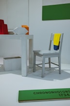 The image features a minimalist installation with a light gray chair draped with a yellow and blue cloth. A white, rectangular table is present, with geometric shapes including a red object, an orange cube, and a white polygon. A green and white two-toned wall decoration is visible, along with a green plank on the floor with black text 'CHRONONHOTONTHO 1734'. The setting appears to be an art or design exhibition.
