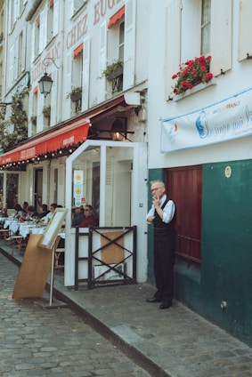 A quaint street scene featuring a French restaurant with a white and red exterior. Tables are set up outside under a canopy with people dining. A man in formal attire stands in front of the restaurant, appearing to be a waiter taking a break. The building facade has red flowers in window boxes and a worn cobblestone street runs past.