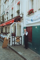 A quaint street scene featuring a French restaurant with a white and red exterior. Tables are set up outside under a canopy with people dining. A man in formal attire stands in front of the restaurant, appearing to be a waiter taking a break. The building facade has red flowers in window boxes and a worn cobblestone street runs past.