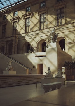 An elegant interior space in a museum, with classical sculptures displayed on raised platforms. Sunlight filters through a glass ceiling, casting intricate shadows on the stone walls and floor. The architecture features arched doorways and large windows, characteristic of historic European design.
