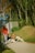 Several people are gathered outdoors on a sunny day. One person is standing with a leash, while others are sitting on the ground, interacting with two playful dogs. The background features a lush, wooded area and a building with a metal facade.