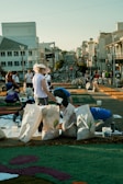A group of people are working on a street art project involving colorful materials spread on the pavement. Large bags filled with different items are placed around them. The background features a city street with several buildings and a clear sky.