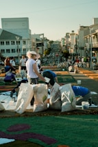 A group of people are working on a street art project involving colorful materials spread on the pavement. Large bags filled with different items are placed around them. The background features a city street with several buildings and a clear sky.