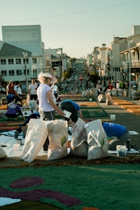 A group of people are working on a street art project involving colorful materials spread on the pavement. Large bags filled with different items are placed around them. The background features a city street with several buildings and a clear sky.