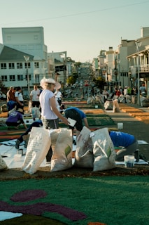 A group of people are working on a street art project involving colorful materials spread on the pavement. Large bags filled with different items are placed around them. The background features a city street with several buildings and a clear sky.
