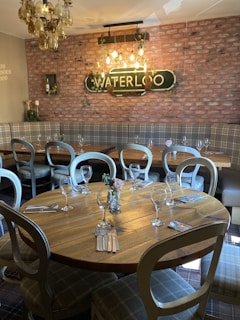 A cozy restaurant interior with a round wooden table set for dining. The table is surrounded by chairs and adorned with an arrangement of wine glasses, cutlery, and a small flower centerpiece. In the background, a brick accent wall features a vintage-style 'Waterloo' sign with hanging light bulbs that illuminate the space. A mix of plaid upholstered seating complements the rustic and inviting atmosphere. A unique chandelier with glass decorations hangs from the ceiling.
