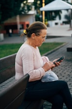 An older woman sits on a park bench, focused on her smartphone. She is wearing a light pink sweater and has her hair pulled back with a yellow hair clip. She holds a plastic bag in her lap.