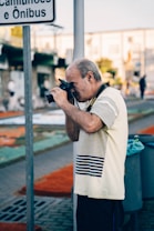 A man in a striped shirt is using a camera to take a photograph in an urban setting. The background features a street with blurred buildings and people, suggesting a busy environment. A signpost and a waste bin are also visible.