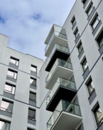 A modern, multi-story residential building with several glass balconies protruding from its facade. The building exterior is mostly composed of light gray concrete with dark gray accents. Windows are evenly spaced across the structure, reflecting the sky above.