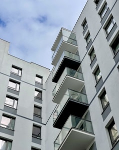 A modern, multi-story residential building with several glass balconies protruding from its facade. The building exterior is mostly composed of light gray concrete with dark gray accents. Windows are evenly spaced across the structure, reflecting the sky above.
