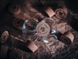 Several glass jars with cork lids are placed on crumpled brown paper. The cork lids have a logo and text printed on them, and some are positioned beside the jars.