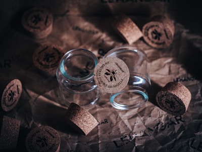 Several glass jars with cork lids are placed on crumpled brown paper. The cork lids have a logo and text printed on them, and some are positioned beside the jars.