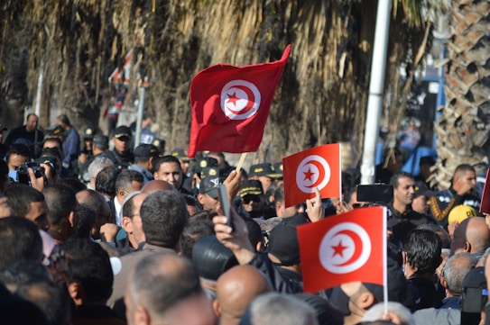 A large crowd gathers outdoors, many holding red and white flags with a crescent and star emblem. People appear engaged in a public demonstration or rally, creating a bustling and energetic atmosphere. Among the crowd, some are holding signs or objects aloft while wearing casual clothing.