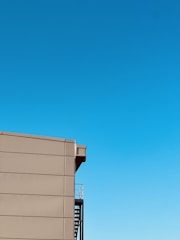 A minimalist image featuring a section of a modern building with a smooth, beige facade and an external staircase. The sky is clear and vibrant blue, occupying the majority of the frame.