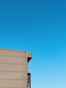 A minimalist image featuring a section of a modern building with a smooth, beige facade and an external staircase. The sky is clear and vibrant blue, occupying the majority of the frame.
