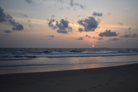 The image features a serene beach setting at sunset, with gentle waves lapping against the shore. The sun is partially visible on the horizon, casting a warm glow across the sky, which is adorned with scattered clouds. The sand appears smooth with faint footprints leading away from the viewer.