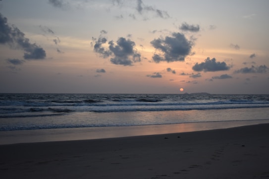 The image features a serene beach setting at sunset, with gentle waves lapping against the shore. The sun is partially visible on the horizon, casting a warm glow across the sky, which is adorned with scattered clouds. The sand appears smooth with faint footprints leading away from the viewer.