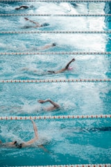 Several swimmers are competing in a pool, performing freestyle strokes as they create splashes in the water. The lanes are clearly marked with orange and white lane dividers, and the water appears vibrant and turquoise.