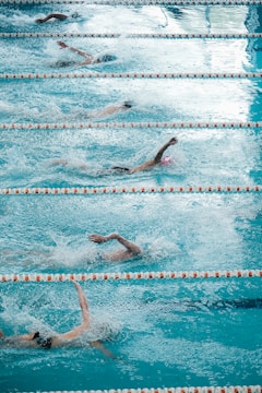 Several swimmers are competing in a pool, performing freestyle strokes as they create splashes in the water. The lanes are clearly marked with orange and white lane dividers, and the water appears vibrant and turquoise.