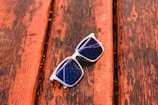 A pair of sunglasses with transparent frames rests on a weathered wooden surface. The lenses appear dark and have a reflective quality, with visible light glare. The wooden surface is painted in a reddish-brown color and shows signs of wear and aging.